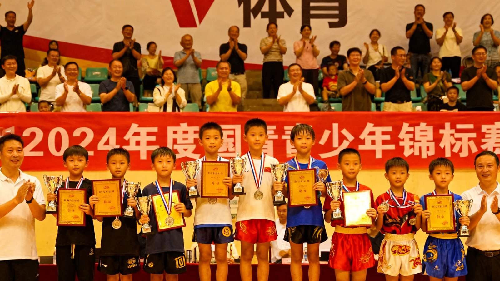 2024年度青少年锦标赛颁奖仪式现场，获奖小选手们手持奖牌和证书在领奖台上合影，背景是V体育品牌横幅，观众席上家长们鼓掌欢呼